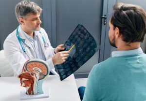 A doctor holding brain scans while explaining to a young male patient what it could mean if he has a headache after a car accident.