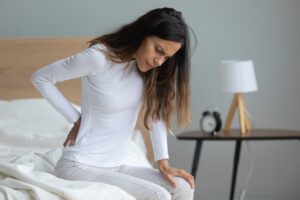 A woman experiencing back pain sits on the edge of her bed and wonders what the common spinal injuries from a car accident are. 