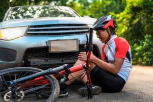 An injured bicyclist sits in front of a car after a crash, waiting to find out what happens if a bicyclist causes an accident.